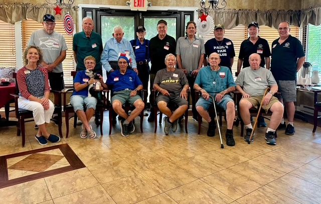 A group of Autumn Ridge residents, several in military-themed hats and shirts, gathered in a common room
